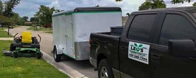 A black truck connected to a trailer. The truck has a Leo Garden Care logo on the side. Beside the truck and trailer is a large zero turn lawn mower.